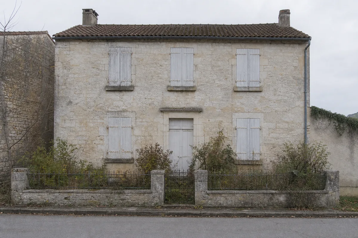 Façade en pierre d’une maison de ville avec volets clos et jardin nu, ciel gris clair